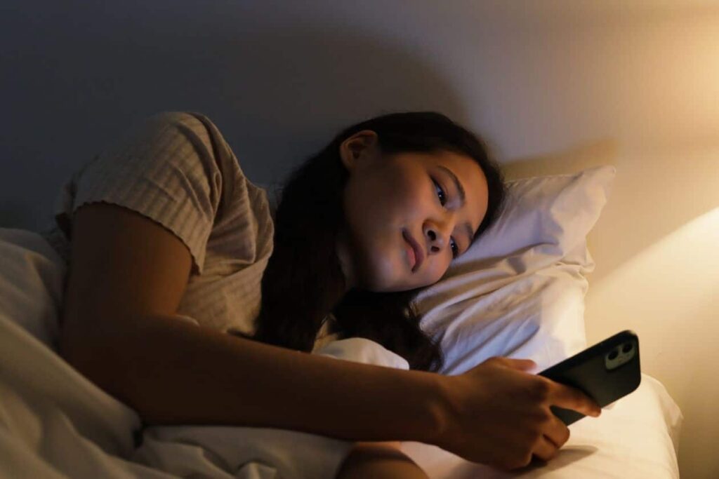Mujer recostada en la cama de noche mirando el teléfono, iluminada por la pantalla