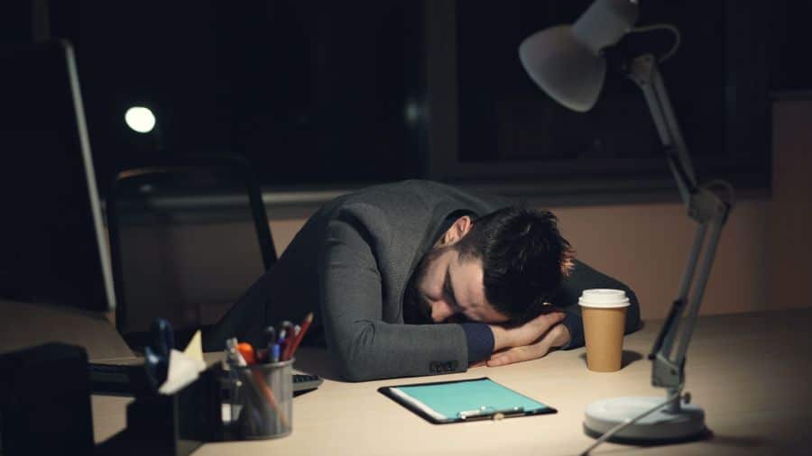 Persona agotada dormida sobre su escritorio de noche, con una taza de café al lado
